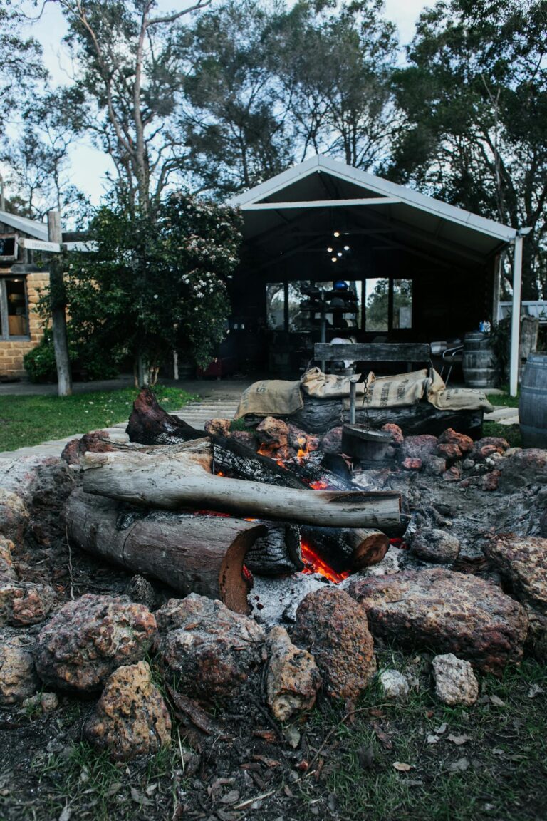 Stone fire pit with burning firewood and charcoal placed on grassy courtyard of residential house with tall trees in countryside